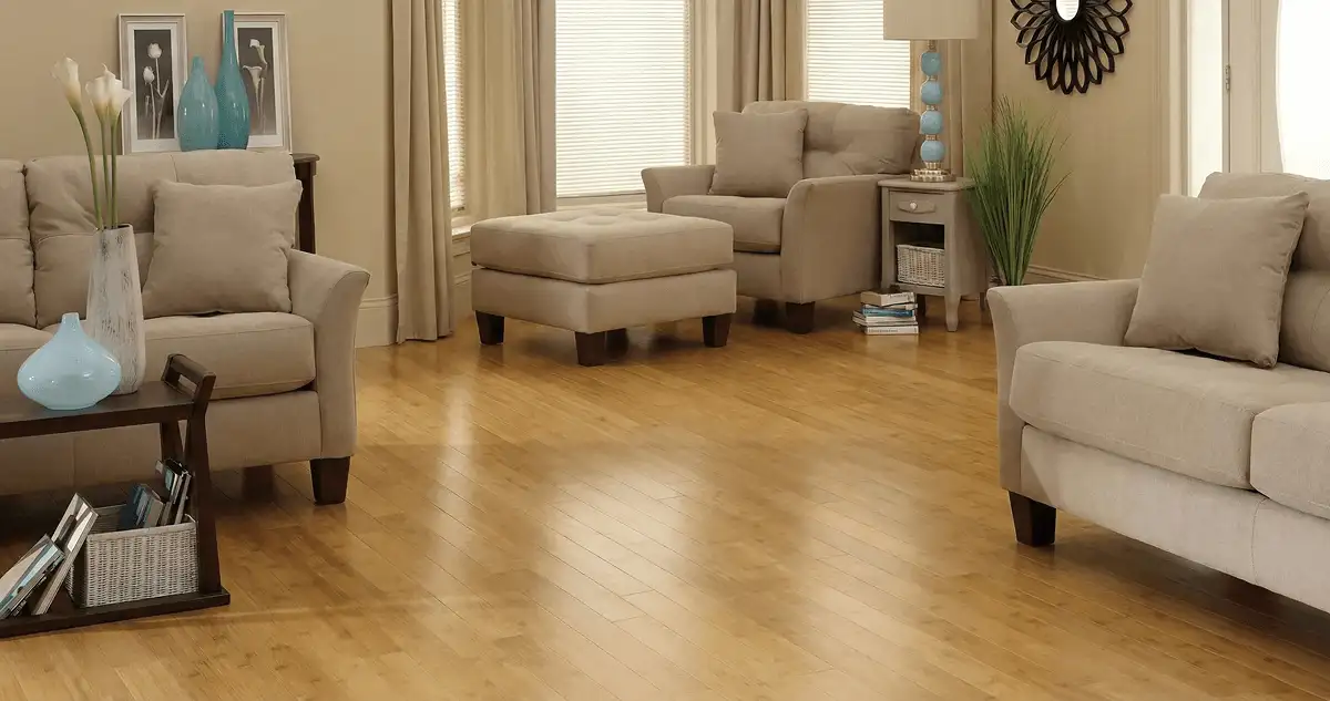A living room with professionally installed bamboo flooring, beige sofas, a matching ottoman, a side table with a blue lamp, vases, framed pictures, and large windows with blinds and curtains in Columbia, MO